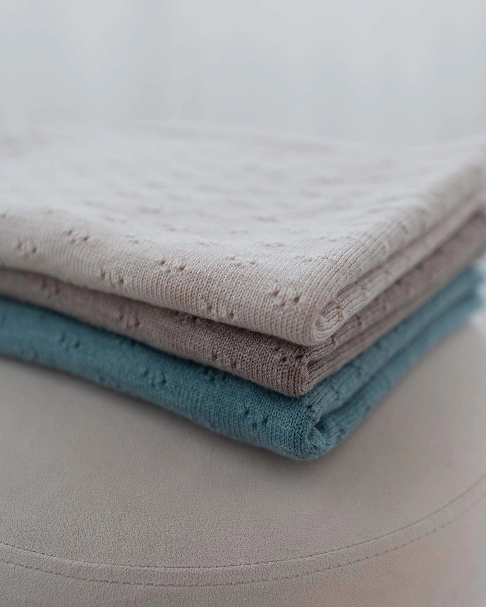 Folded stack of three soft merino wool baby blankets in turquoise, beige, and light gray with delicate eyelet patterns.