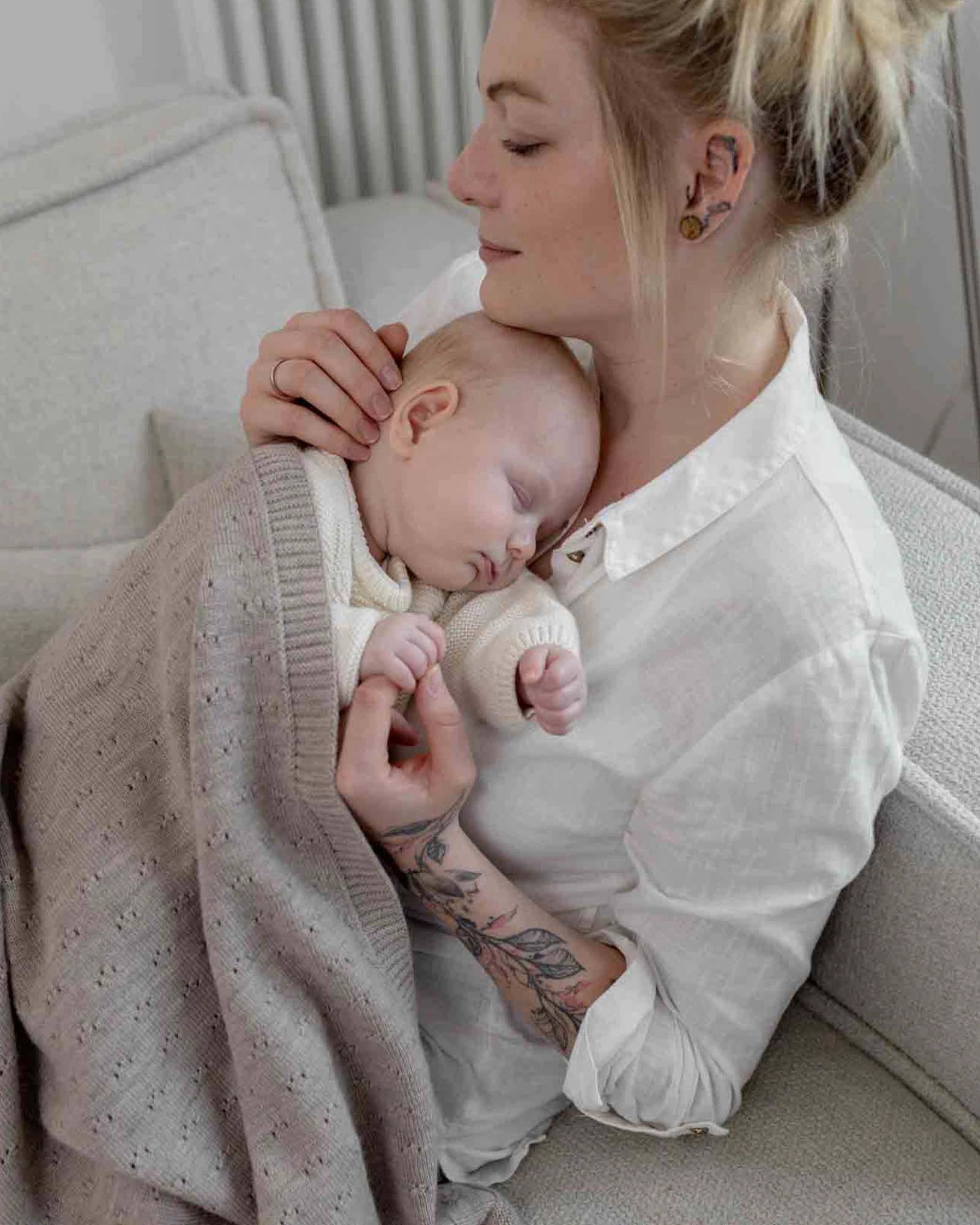 Mother holding sleeping baby wrapped in a soft beige Merino wool blanket with a subtle knit pattern.