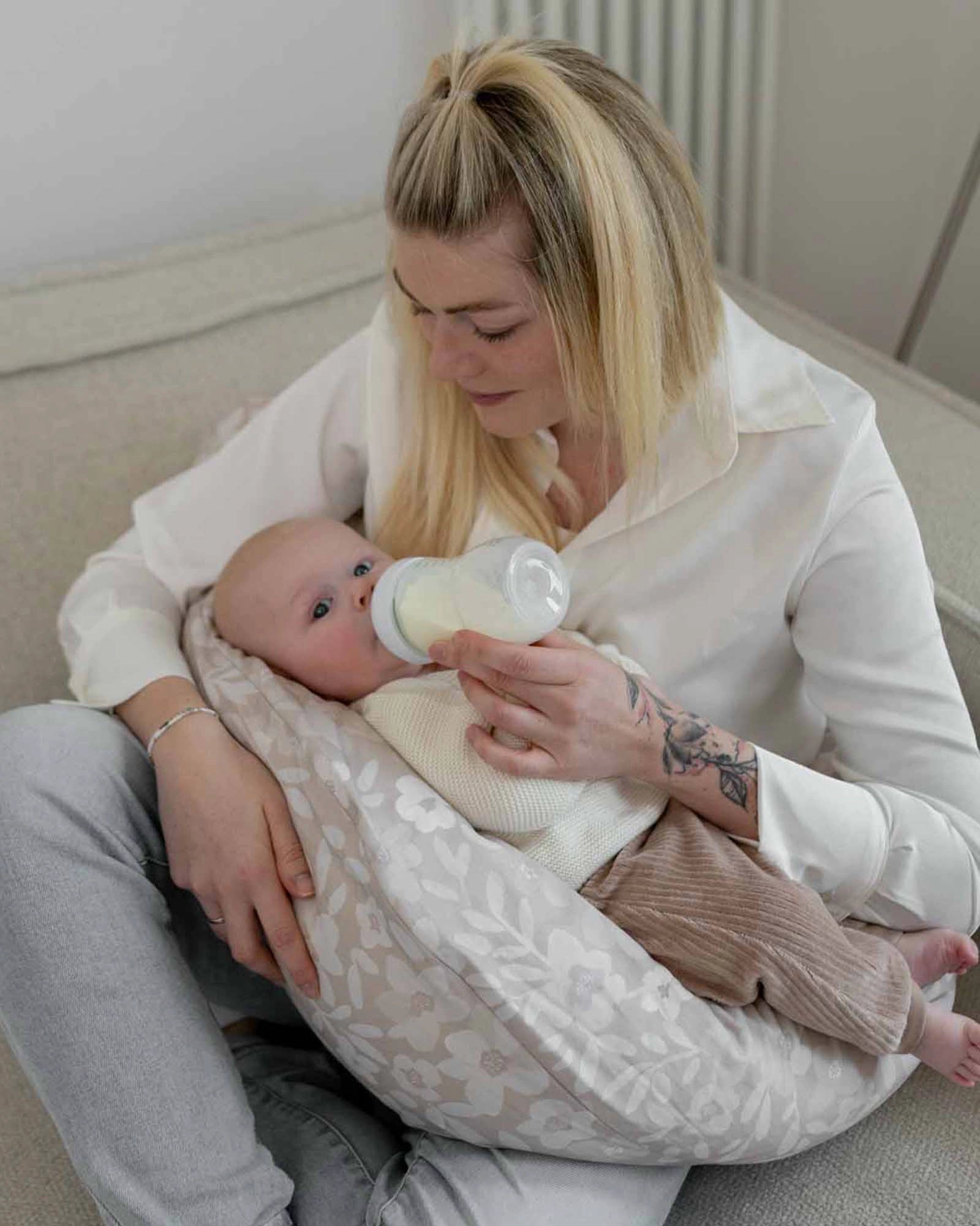 Beige cotton nursing pillow cover with hand-painted floral design, supporting a baby being bottle-fed.