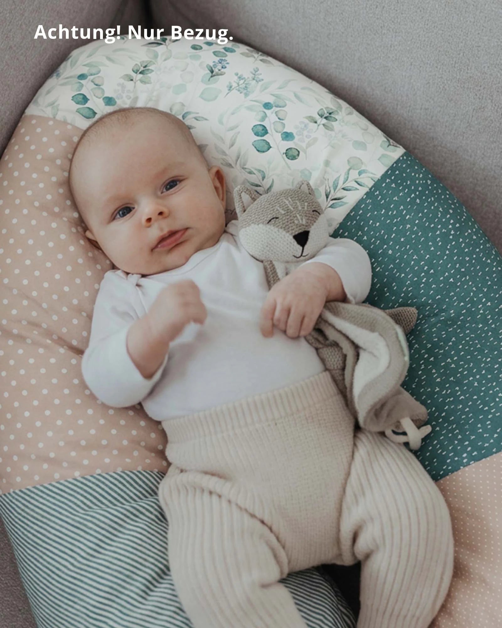 Baby liegt auf einem Stillkissenbezug mit Eukalyptus- und Punktemuster in Grün, Rosa und Beige, hält ein Stofftier.