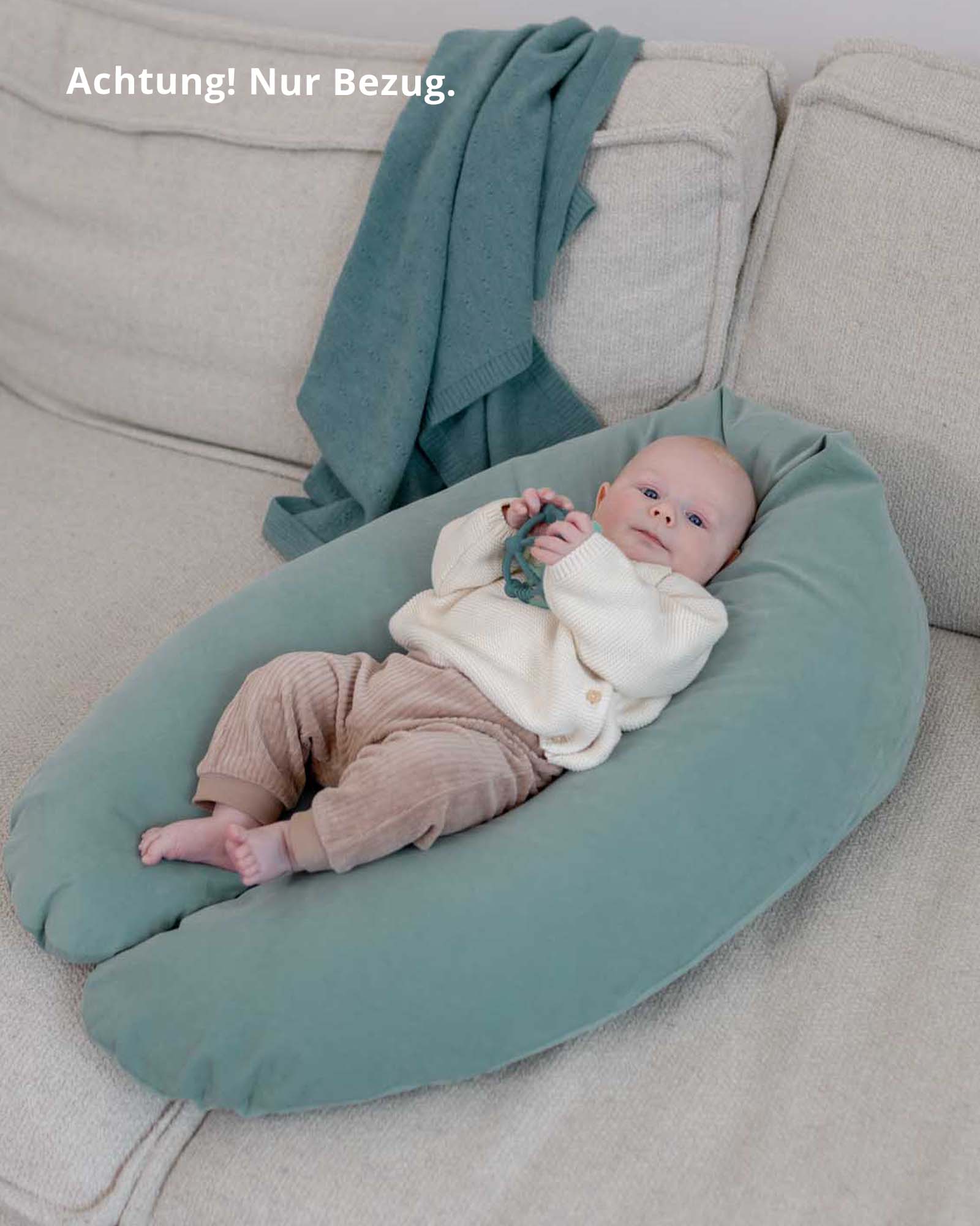 Baby on a sage green corduroy nursing pillow cover, holding a teether, on a light gray sofa.