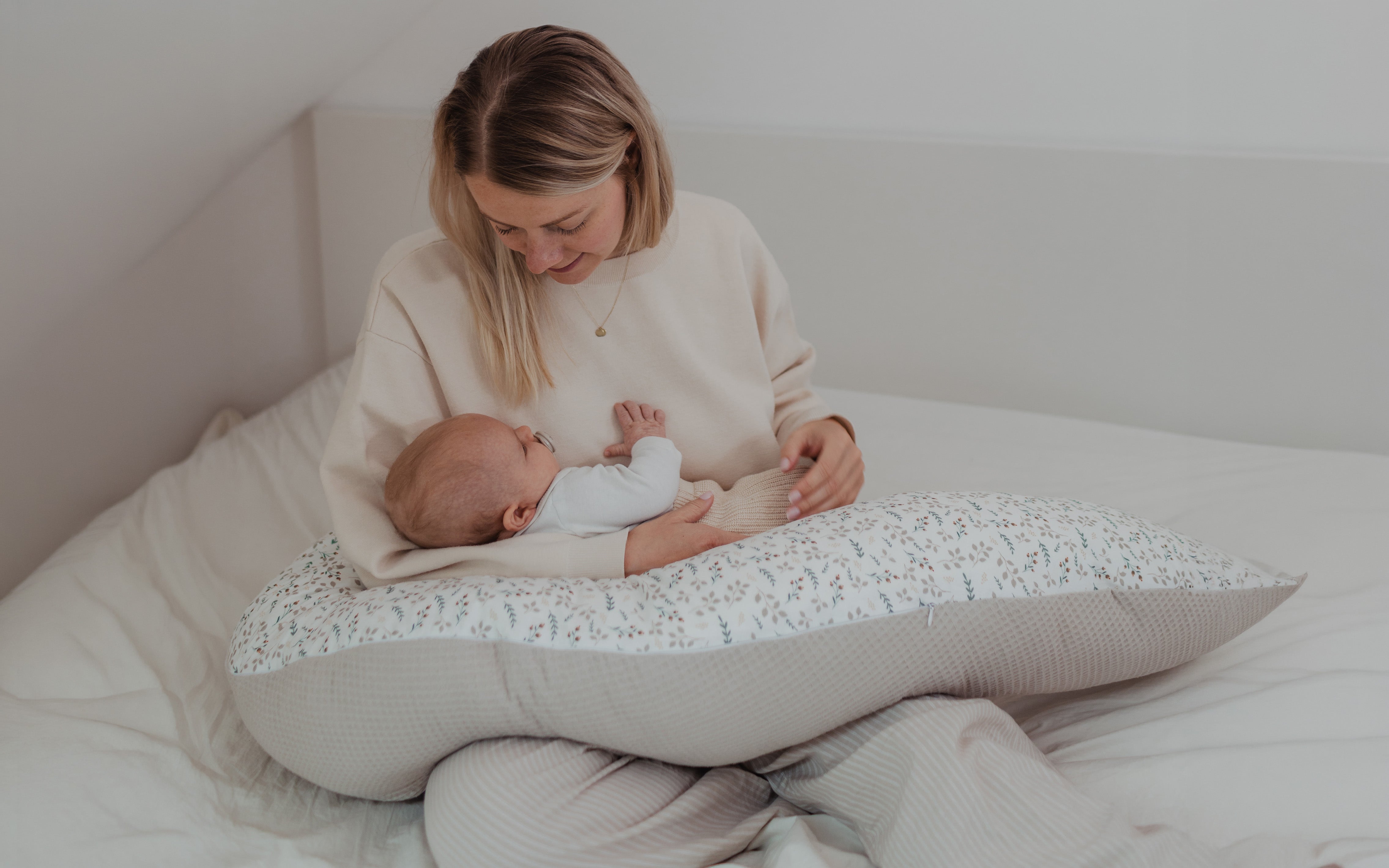 Junge Mutter in beigem Pullover hält Baby auf einem gemusterten Stillkissen mit Blättern und Streifen.