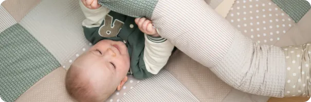 Baby liegt lächelnd auf einer Patchwork-Krabbeldecke und spielt mit einer gemusterten Bettschlange.