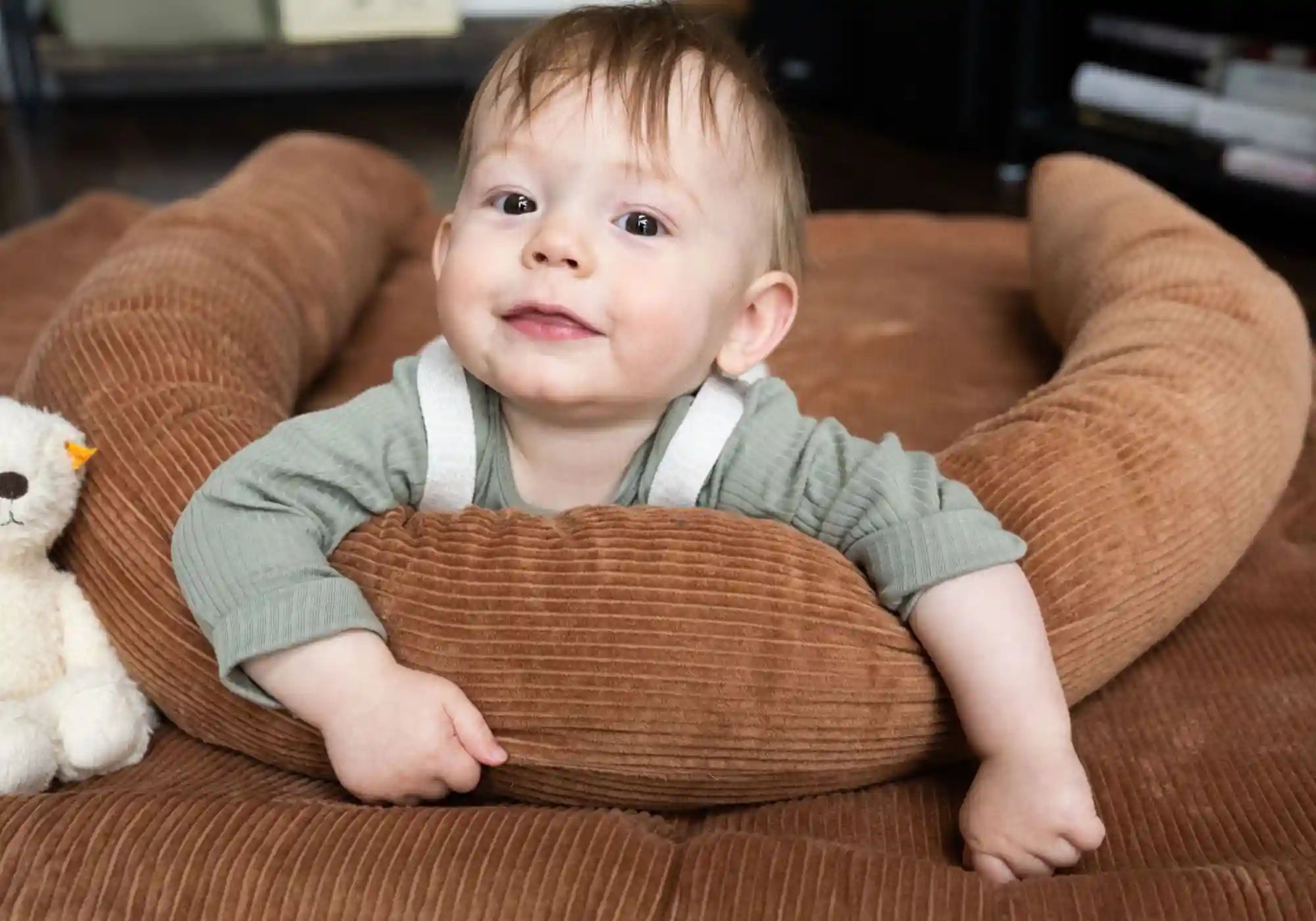 Ein lächelndes Baby liegt in einem U-förmigen, braunen Cord-Stillkissen auf einer passenden Decke neben einem Teddybär.