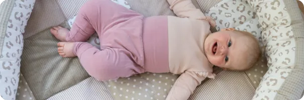 Fröhliches Baby in rosa Kleidung liegt auf einer gepolsterten Krabbeldecke mit Leopardenmuster und Punkten.