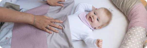 Fröhliches Baby liegt im Bettchen unter einer ULLENBOOM Märchenwald Babydecke mit rosa und beigen Stoffen.