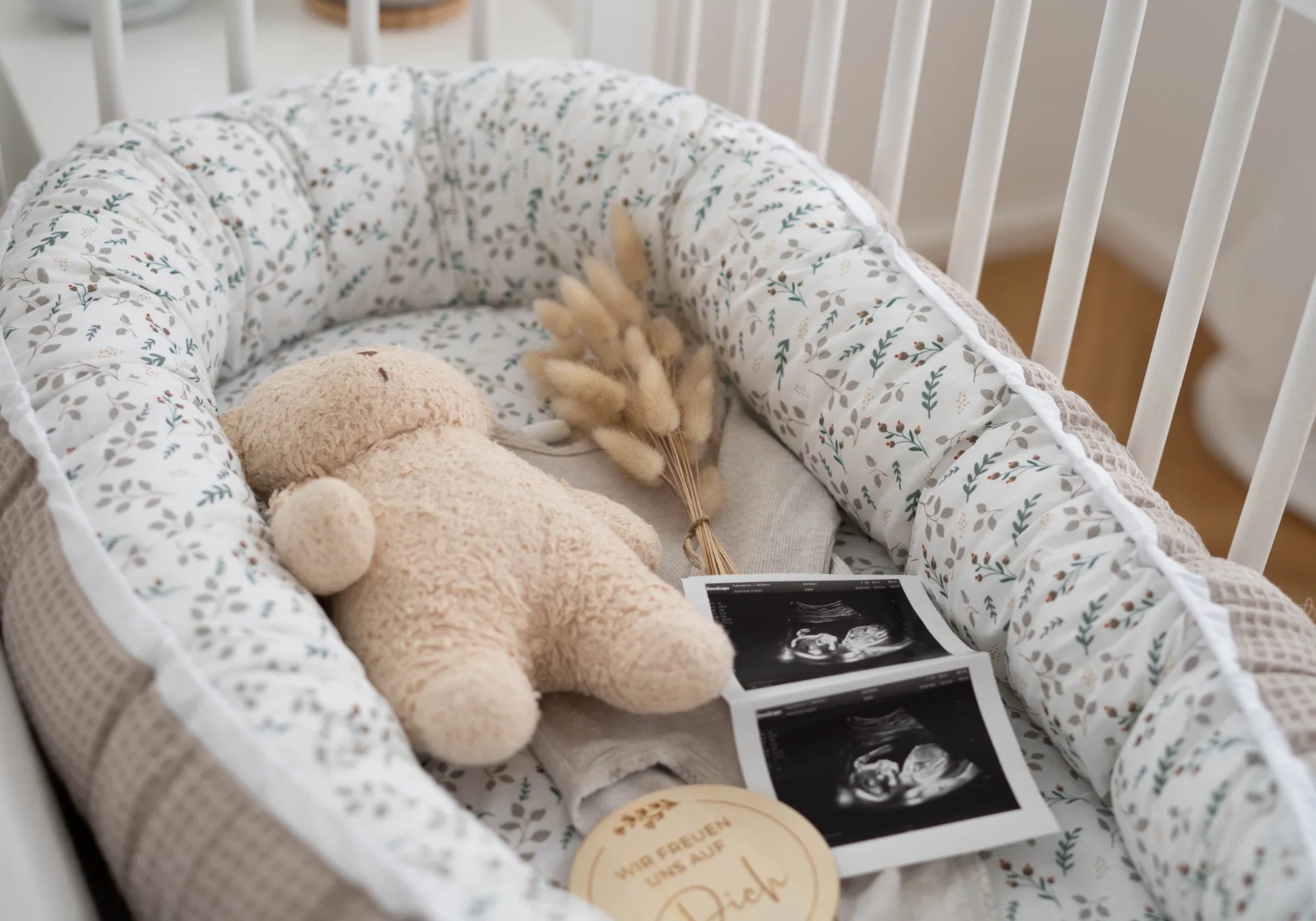 Baby Nestchen mit floralem Sand-Muster, Teddybär, getrockneten Blumen und Ultraschallbildern im Babybett.