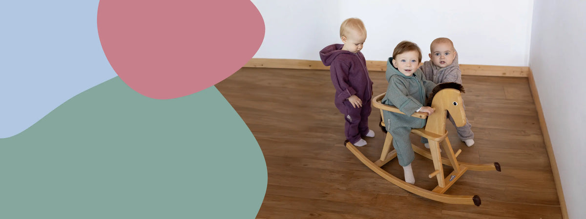Drei Kleinkinder in farbigen Walk-Anzügen von ULLENBOOM spielen gemeinsam auf einem hölzernen Schaukelpferd in einem hellen Raum.