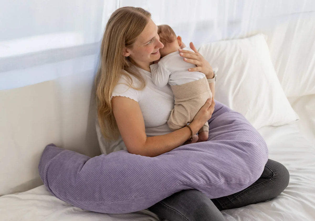 Eine Mutter hält ihr Baby auf einem fliederfarbenen ULLENBOOM Stillkissen aus weichem Cordstoff auf einem weißen Bett.