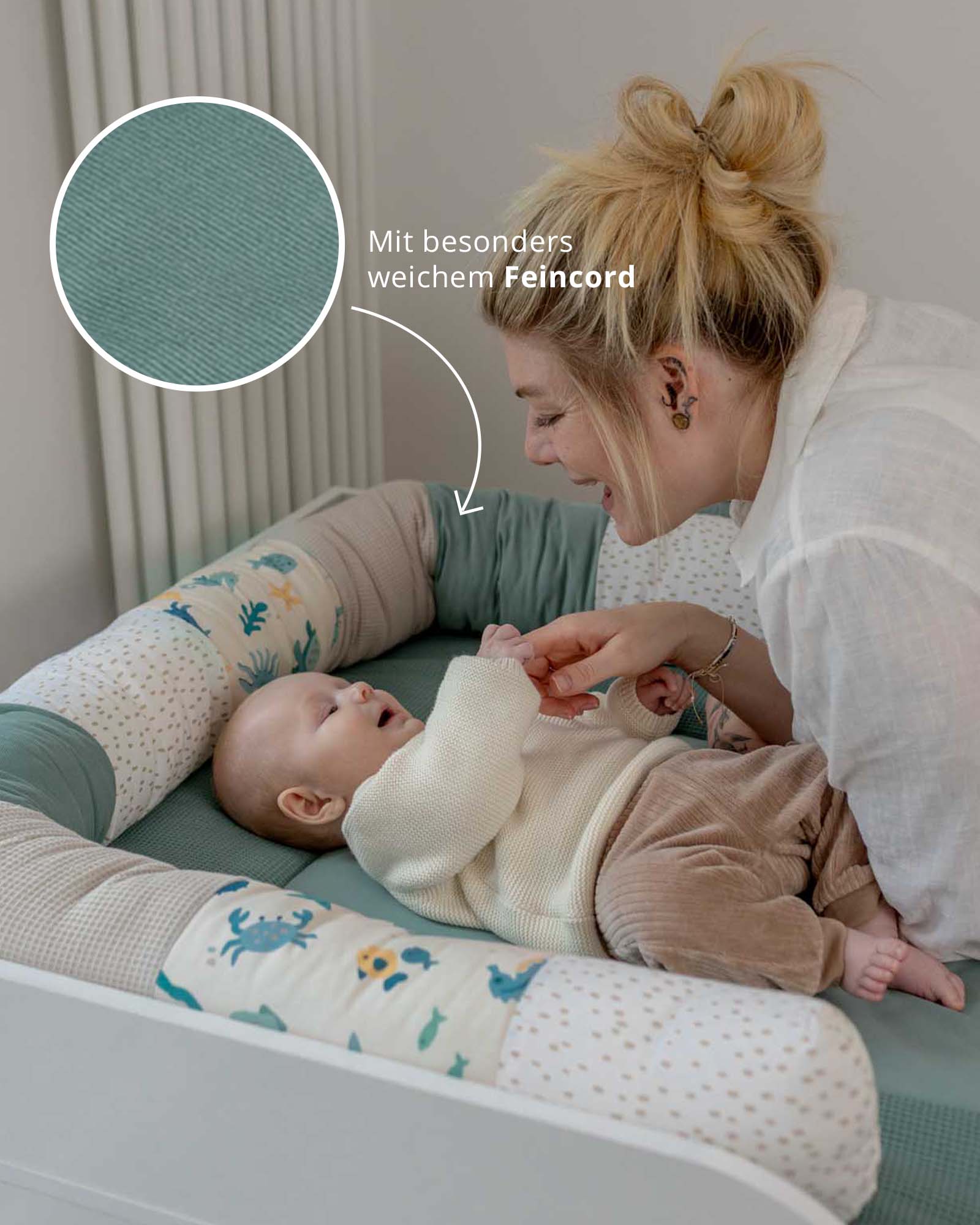 Baby Bettschlange in Unterwasserwelt-Design mit Feincord-Patches, umgibt ein Baby auf einer Wickelkommode.
