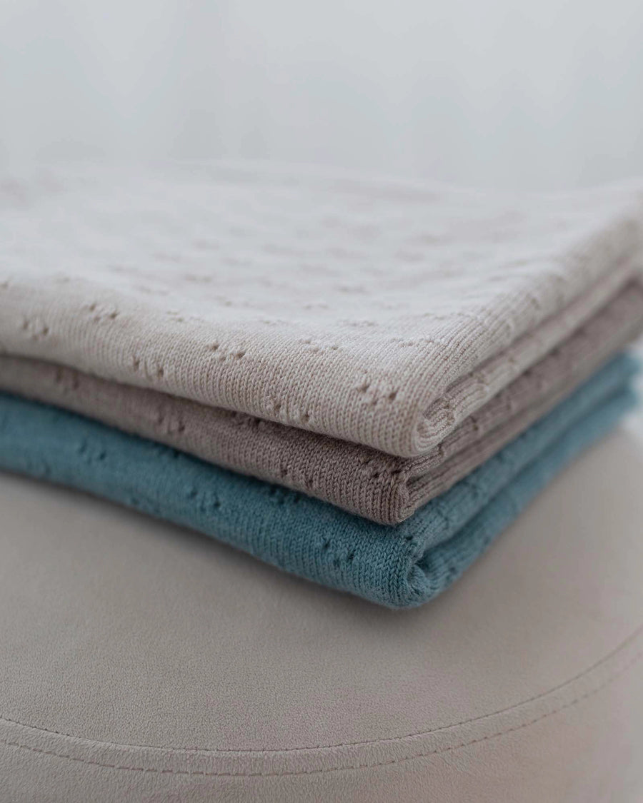 Folded stack of three soft merino wool baby blankets in turquoise, beige, and light gray with delicate eyelet patterns.
