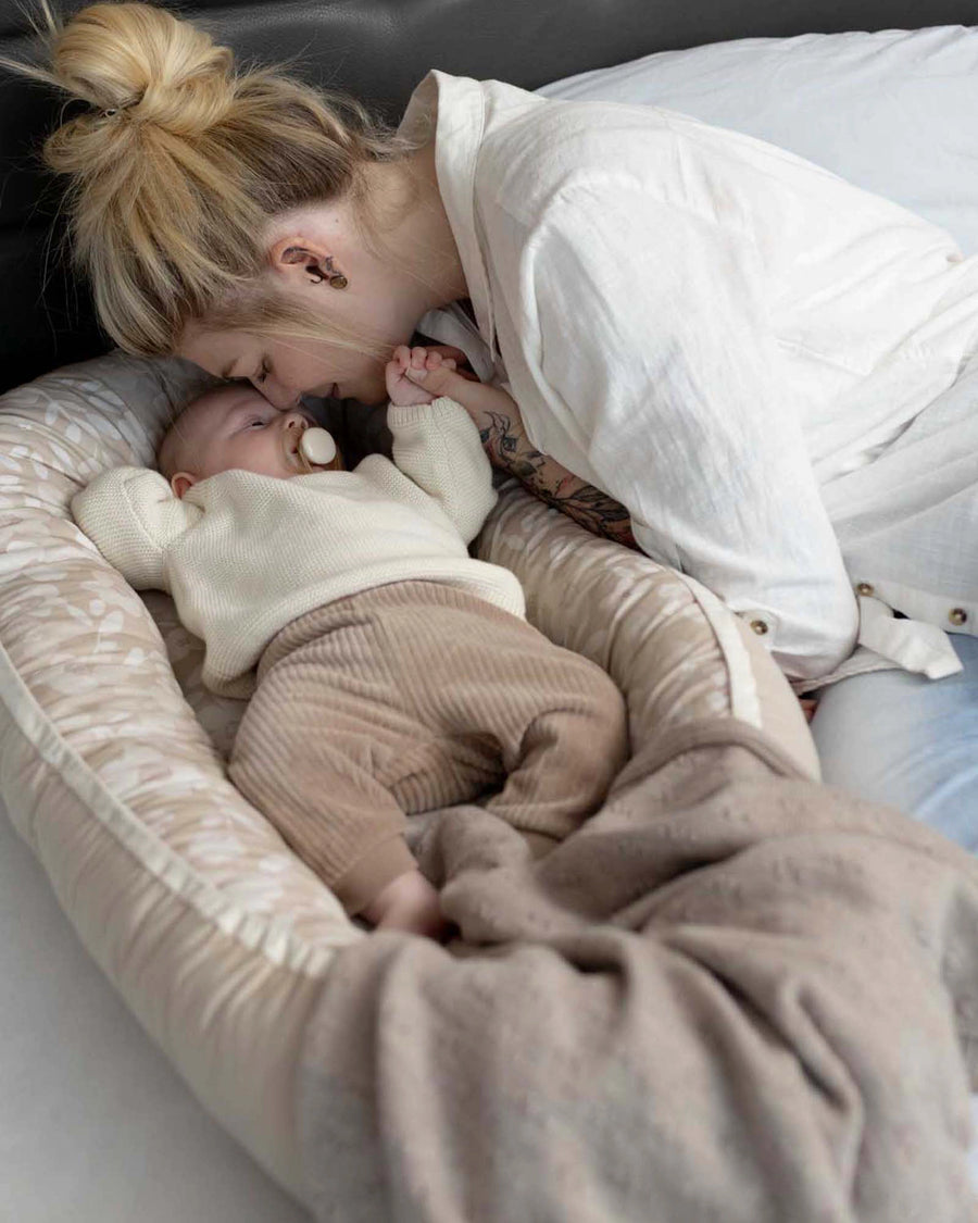 Mother gently kisses baby in a reversible, beige patterned babynest with a soft blanket.