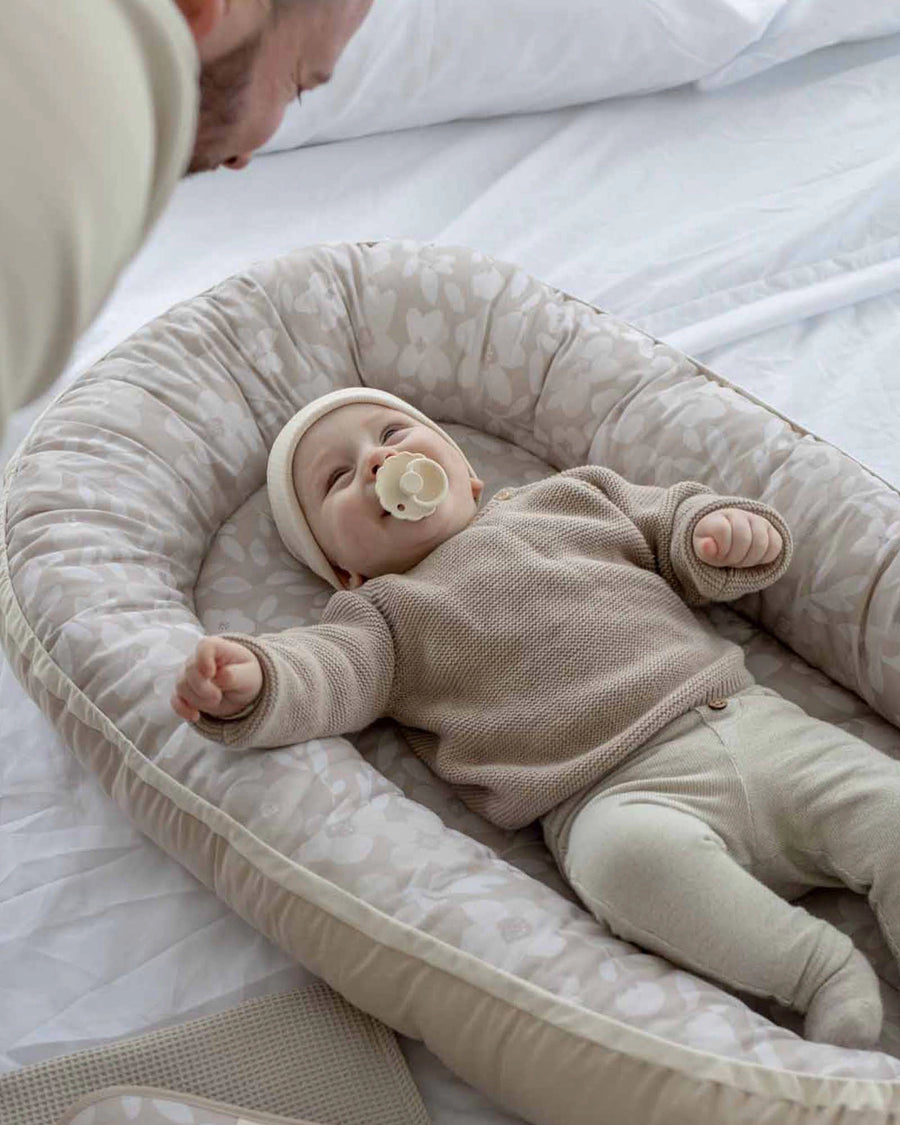 Baby in a beige floral babynest with a pacifier, looking up at a parent.