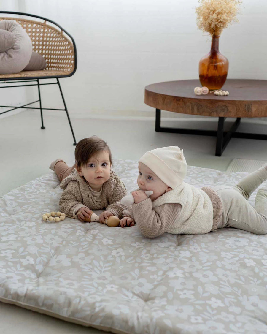 Two babies on a thick, round, beige playmat with a subtle white floral pattern.