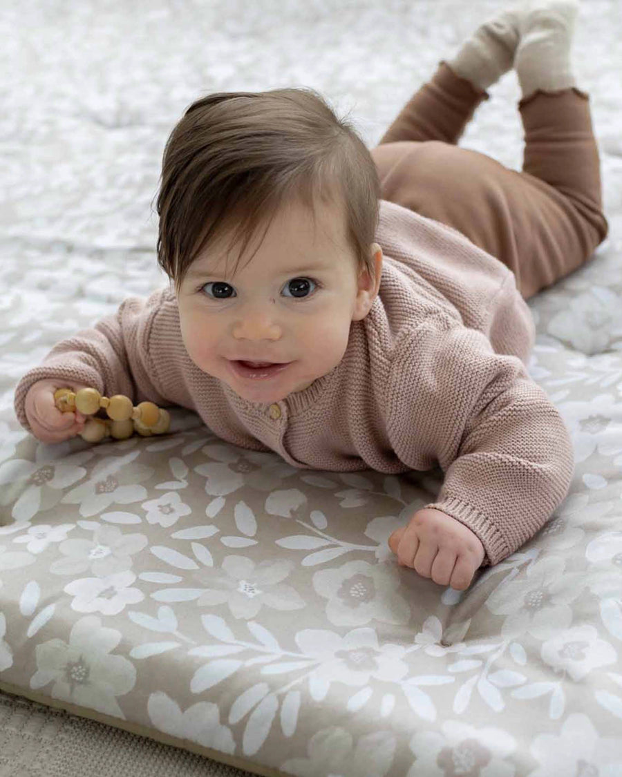 Baby on a thick, soft playmat with hand-painted beige and white floral design, holding a wooden teether.