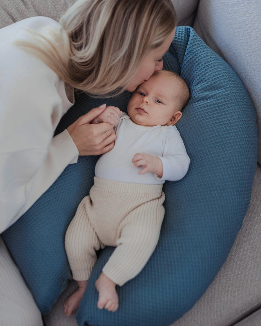 Mutter küsst Baby, das auf einem blauen Stillkissen mit Waffelpiqué-Bezug liegt, ideal für Stillzeit und Schwangerschaft.