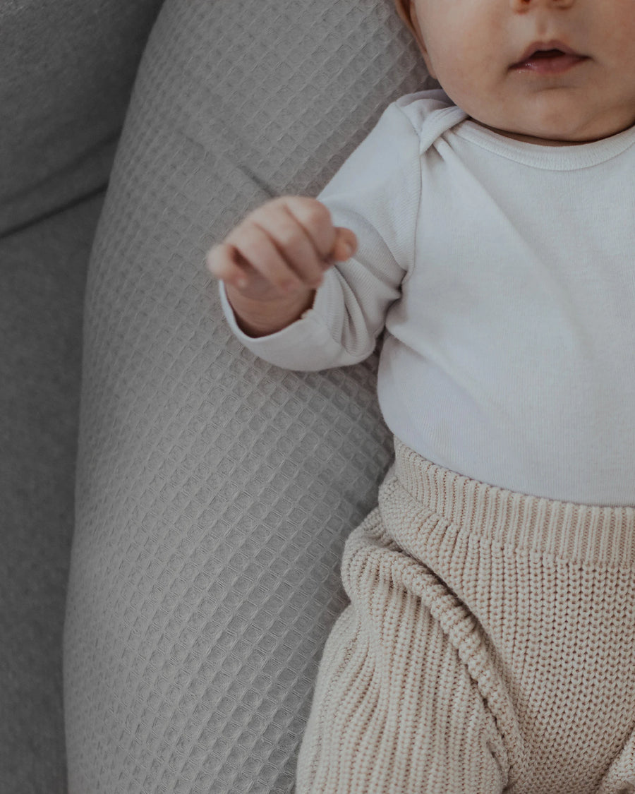 Baby liegt auf einem grauen Stillkissen mit Waffelpiqué-Bezug, trägt einen weißen Body und beige Strickhose.