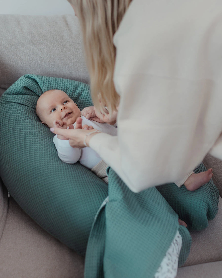 Baby liegt auf einem salbeigrünen Stillkissen aus Waffelpiqué, eine Person berührt sanft sein Gesicht.