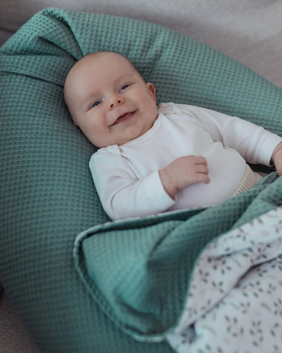 Lächelndes Baby liegt bequem auf einem salbeigrünen Stillkissen aus Waffelpiqué mit einer gemusterten Decke.