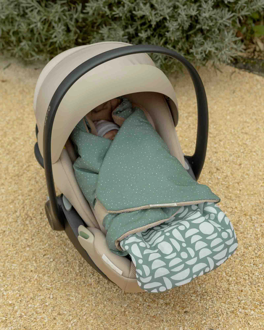 Baby sleeping soundly in a beige car seat, covered by a green Einschlagdecke with white polka dots and a patterned footmuff.