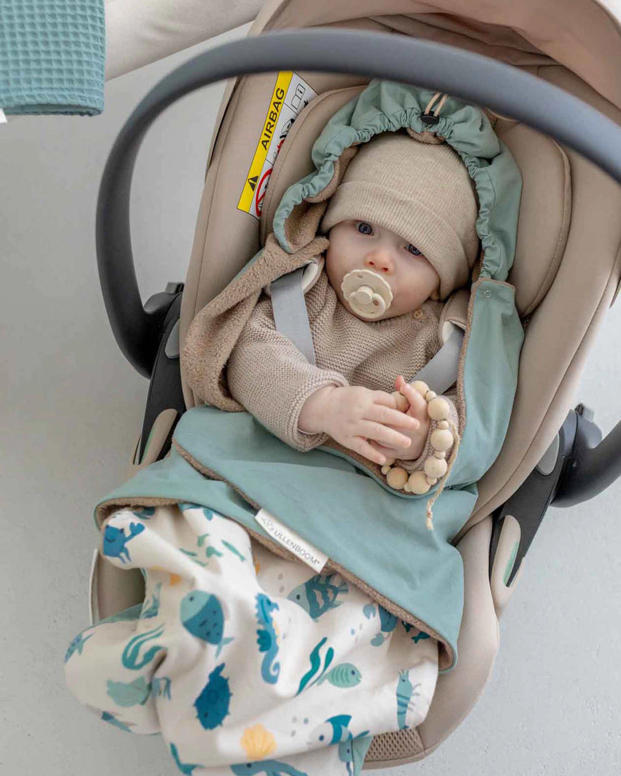 Baby in a beige car seat, wearing a beige hat and sweater, tucked into a reversible blue-green and underwater-themed swaddle blanket.