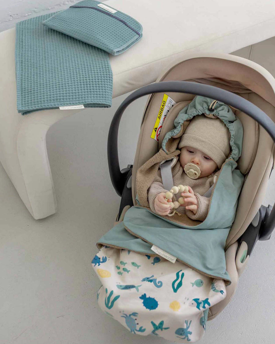 Baby in a beige car seat, wrapped in a blue-green Einschlagdecke blanket with an underwater world pattern.