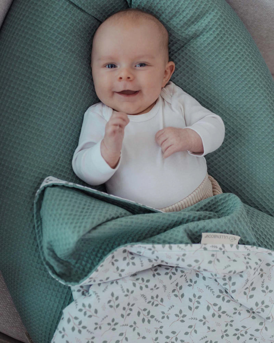Lächelndes Baby in einem grünen Stillkissen mit Waffelstruktur und einer floral gemusterten Decke.