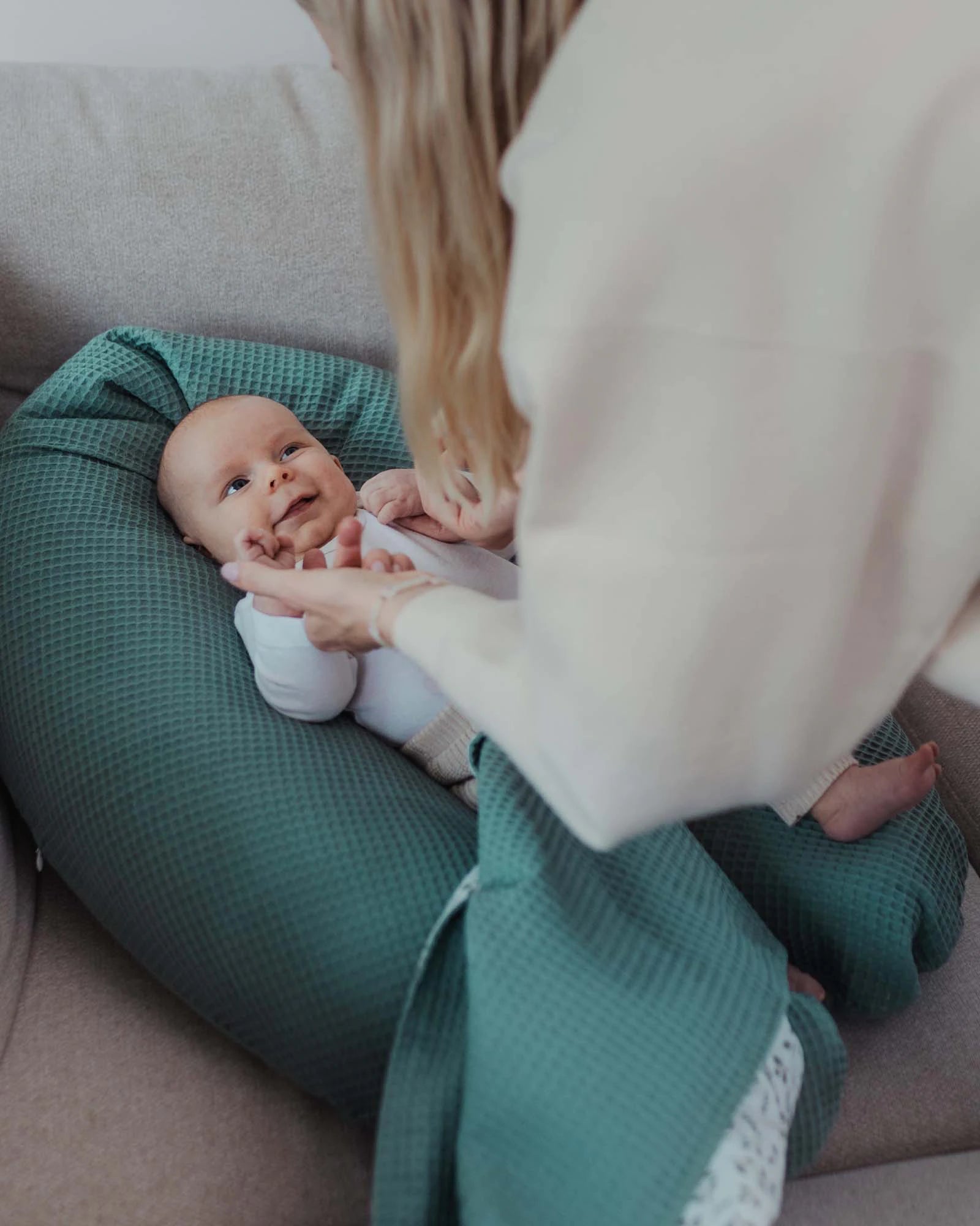 Baby liegt lächelnd auf einem grünen, floral gemusterten Stillkissen aus Baumwolle, eine Frau beugt sich darüber.
