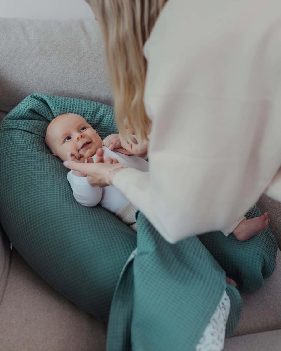 Baby liegt lächelnd auf einem grünen, floral gemusterten Stillkissen aus Baumwolle, eine Frau beugt sich darüber.