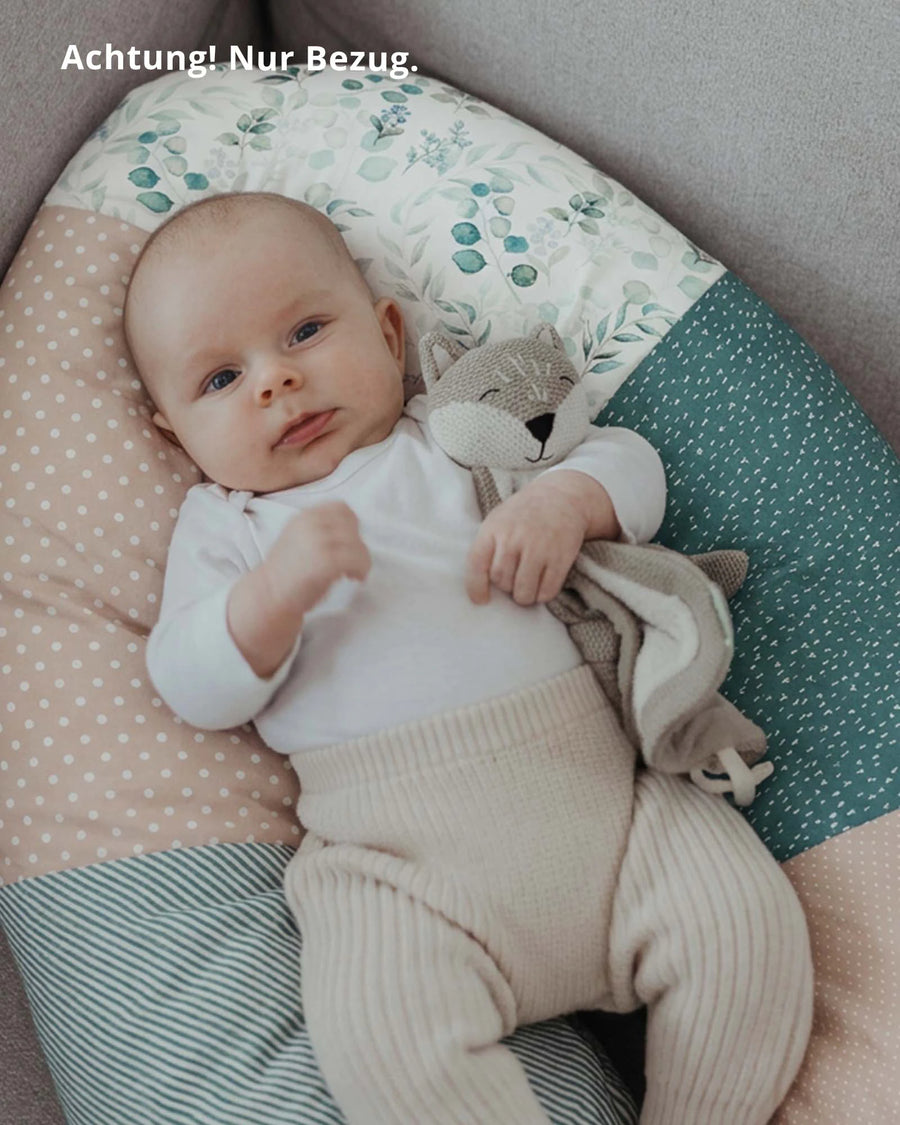 Baby liegt auf einem Stillkissenbezug mit Eukalyptus- und Punktemuster in Grün, Rosa und Beige, hält ein Stofftier.