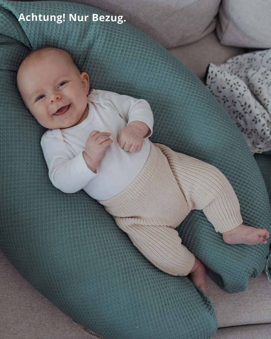 Lachendes Baby liegt auf einem salbeigrünen Stillkissenbezug aus Waffelpiqué, der auf einem Sofa platziert ist.