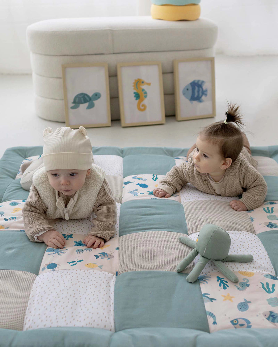 Two babies on a quilted "Underwater World" playmat with sea creatures, a plush octopus, and framed ocean art.