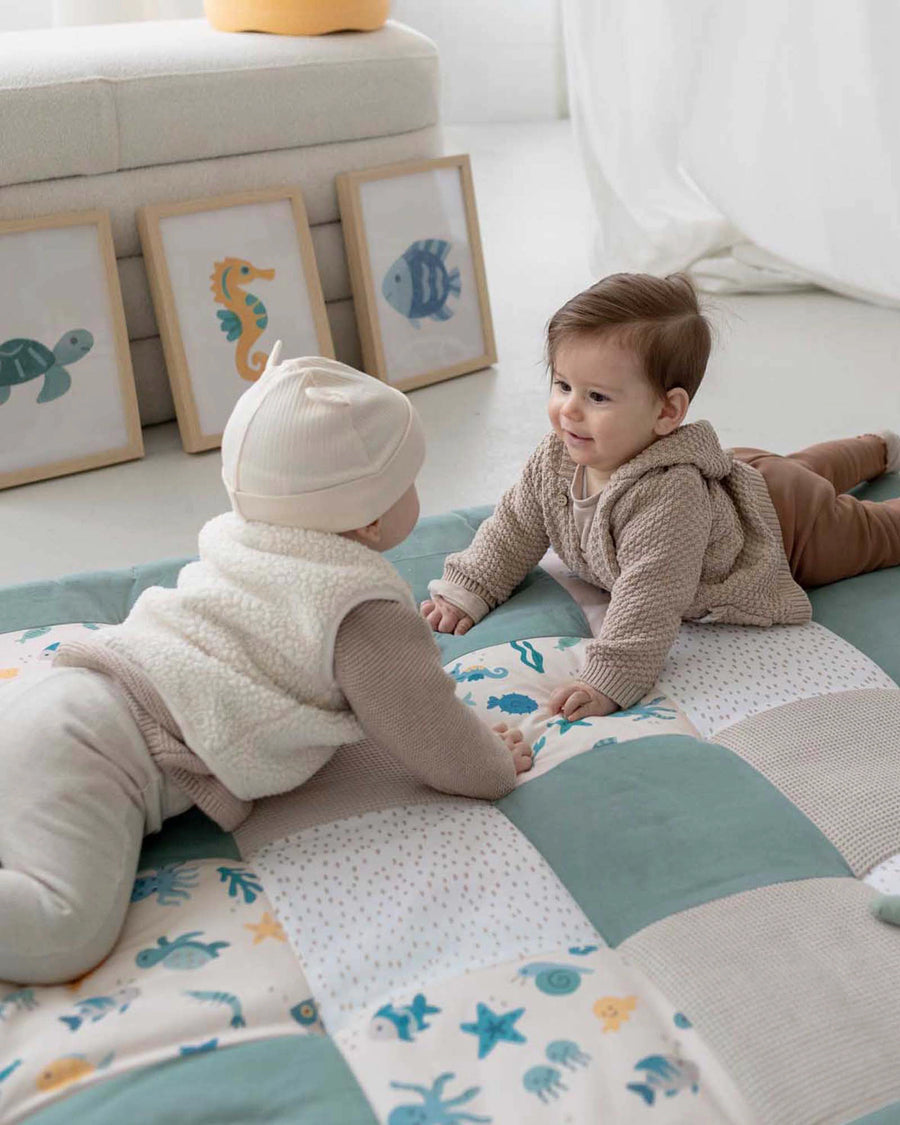 Two babies on a quilted playmat with underwater sea creature patterns in blue, beige, and white.
