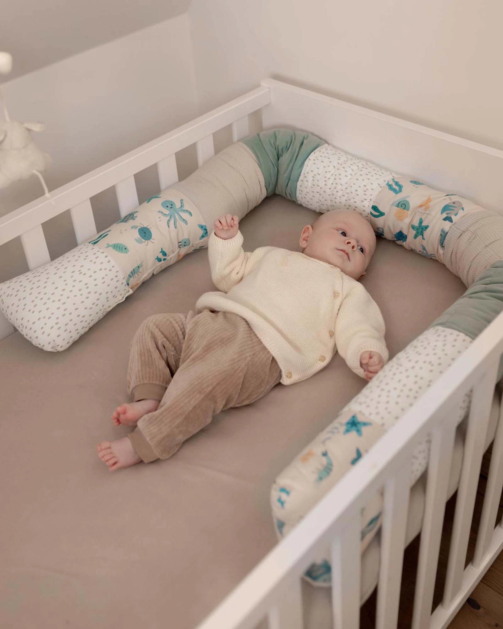 Baby in a white crib with a soft, thick Bettschlange featuring an underwater world design in blue, green, and white.