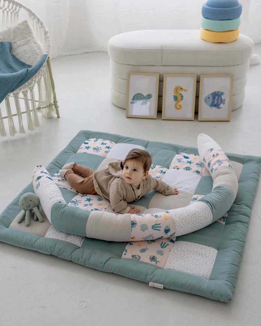 Baby on a soft, quilted playmat with an underwater-themed bettschlange in blue, white, and pink.