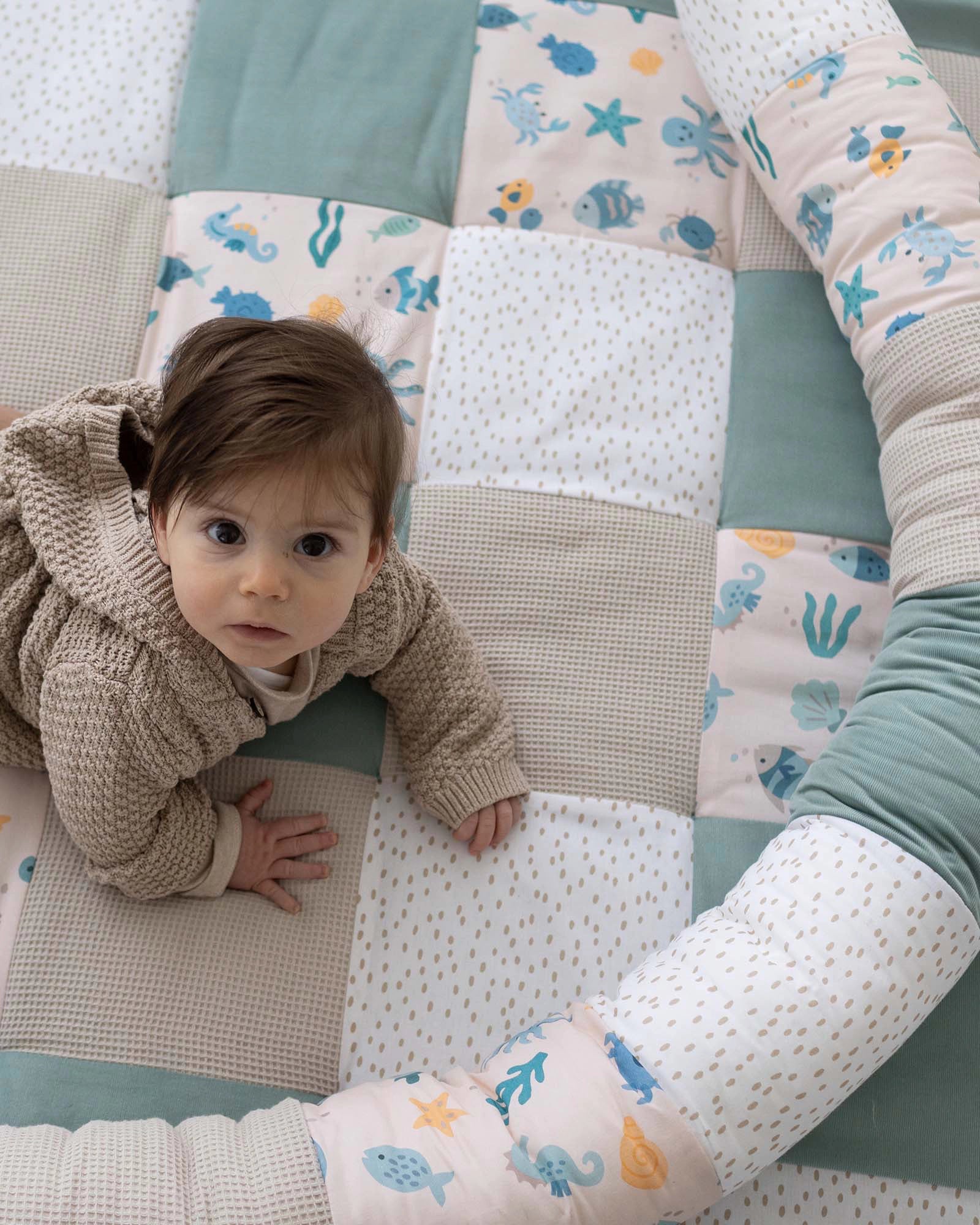 Baby on a quilted playmat with an underwater-themed bed snake in soft blues, pinks, and beige.