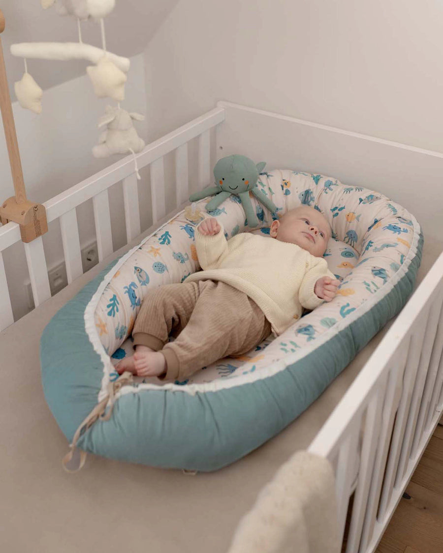 Baby asleep in a reversible Babynest with an underwater world pattern and a teal bottom, inside a white crib.