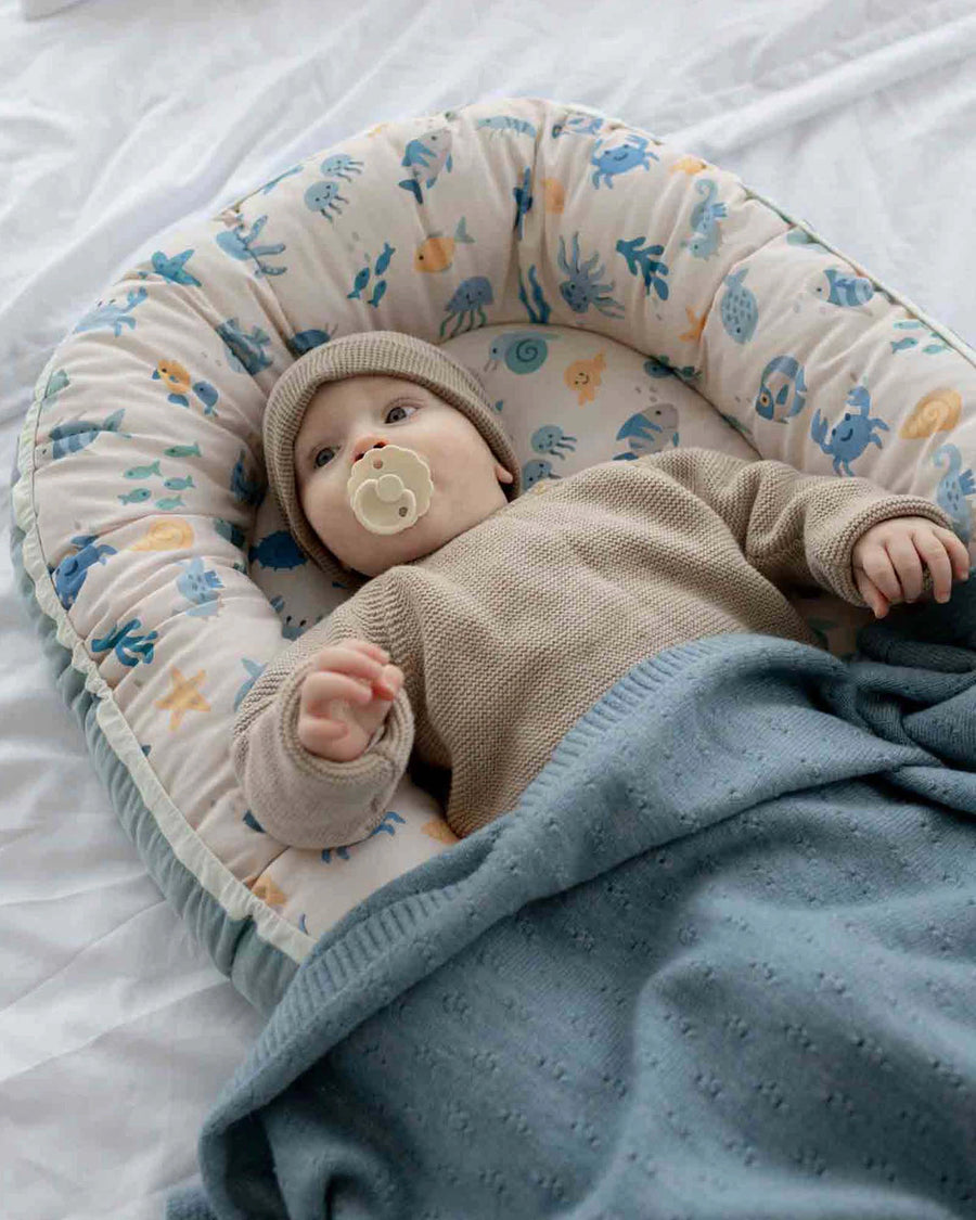 Baby in a reversible Babynest with an underwater sea creature pattern, light blue blanket, and beige knit hat.