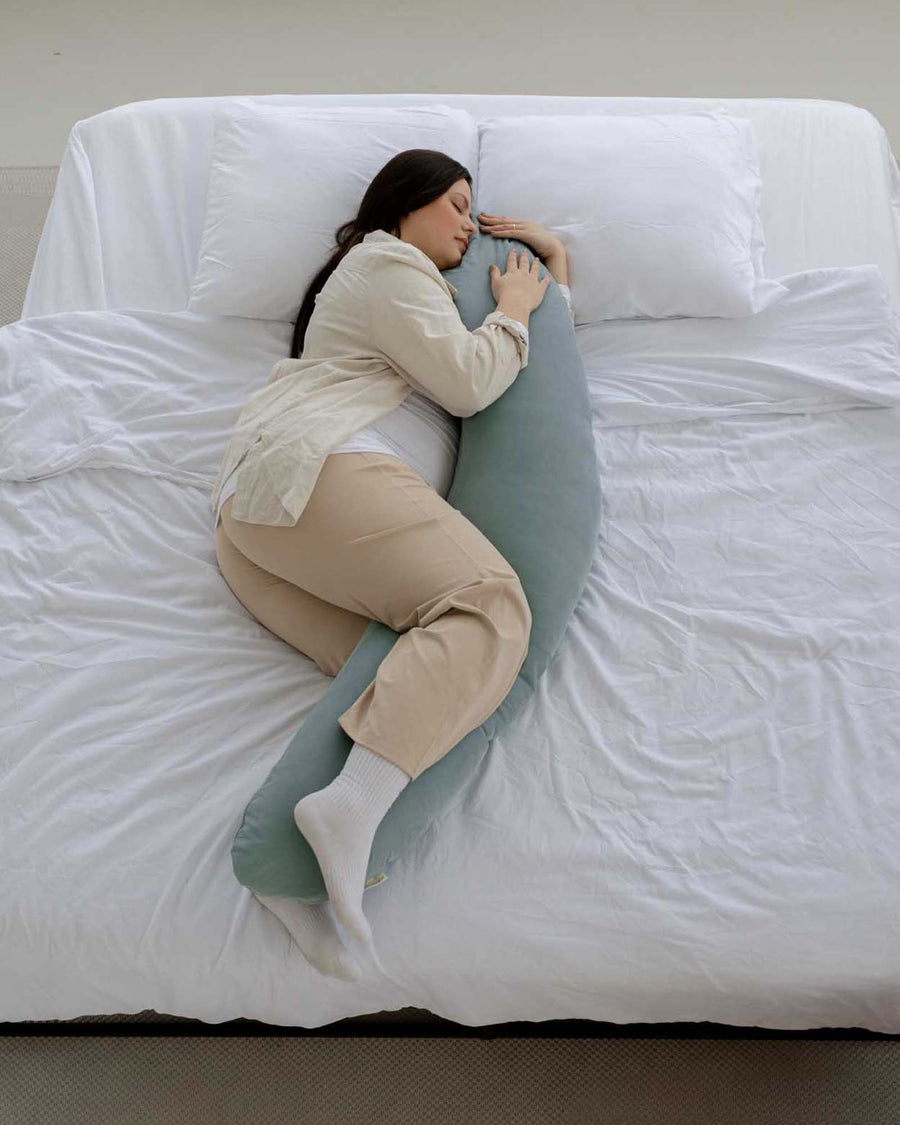 Pregnant woman sleeping on a bed, hugging a soft, light green corduroy nursing pillow.