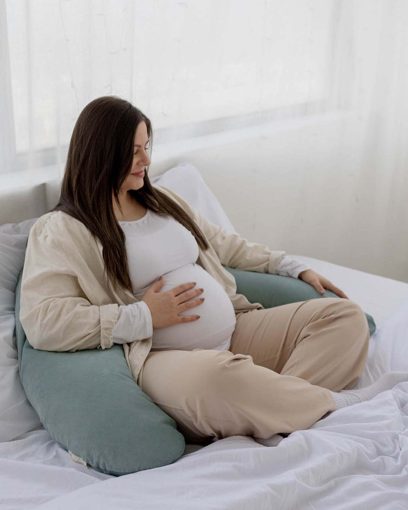 Pregnant woman relaxing on a bed with a light green corduroy U-shaped nursing pillow.