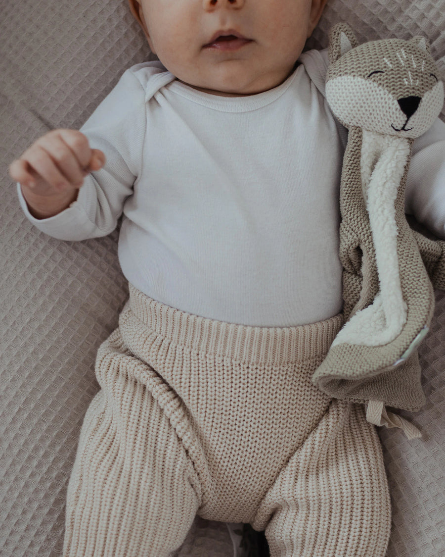 Baby liegt auf einem sandfarbenen Stillkissen mit Waffelpiqué-Bezug und einem gestrickten Fuchs-Kuscheltier.