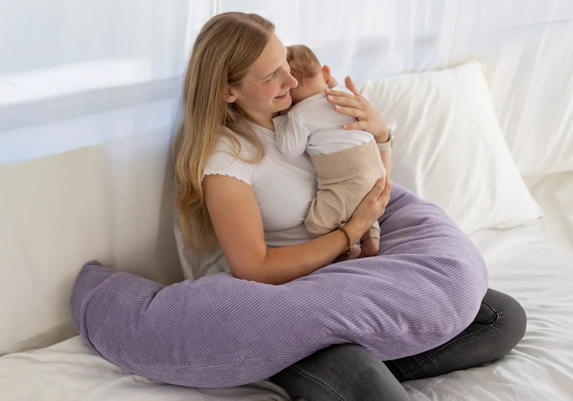 Eine Mutter hält ihr Baby auf einem fliederfarbenen ULLENBOOM Stillkissen aus weichem Cordstoff auf einem weißen Bett.