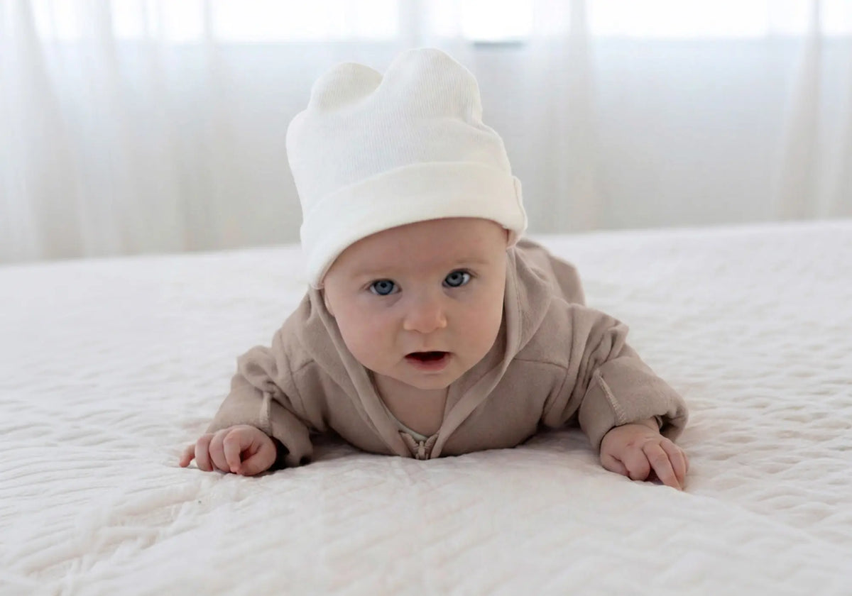Ein Baby in beigem Strampler und weißer Mütze liegt auf dem Bauch auf einer hellen, strukturierten Decke.